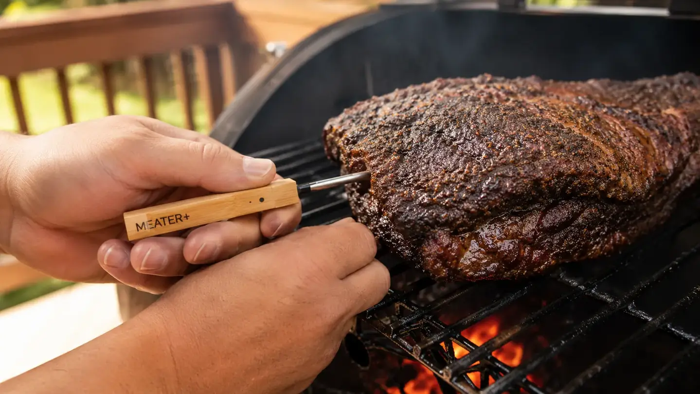 A hand guiding a wireless thermometer probe into a brisket set on a smoker grate
