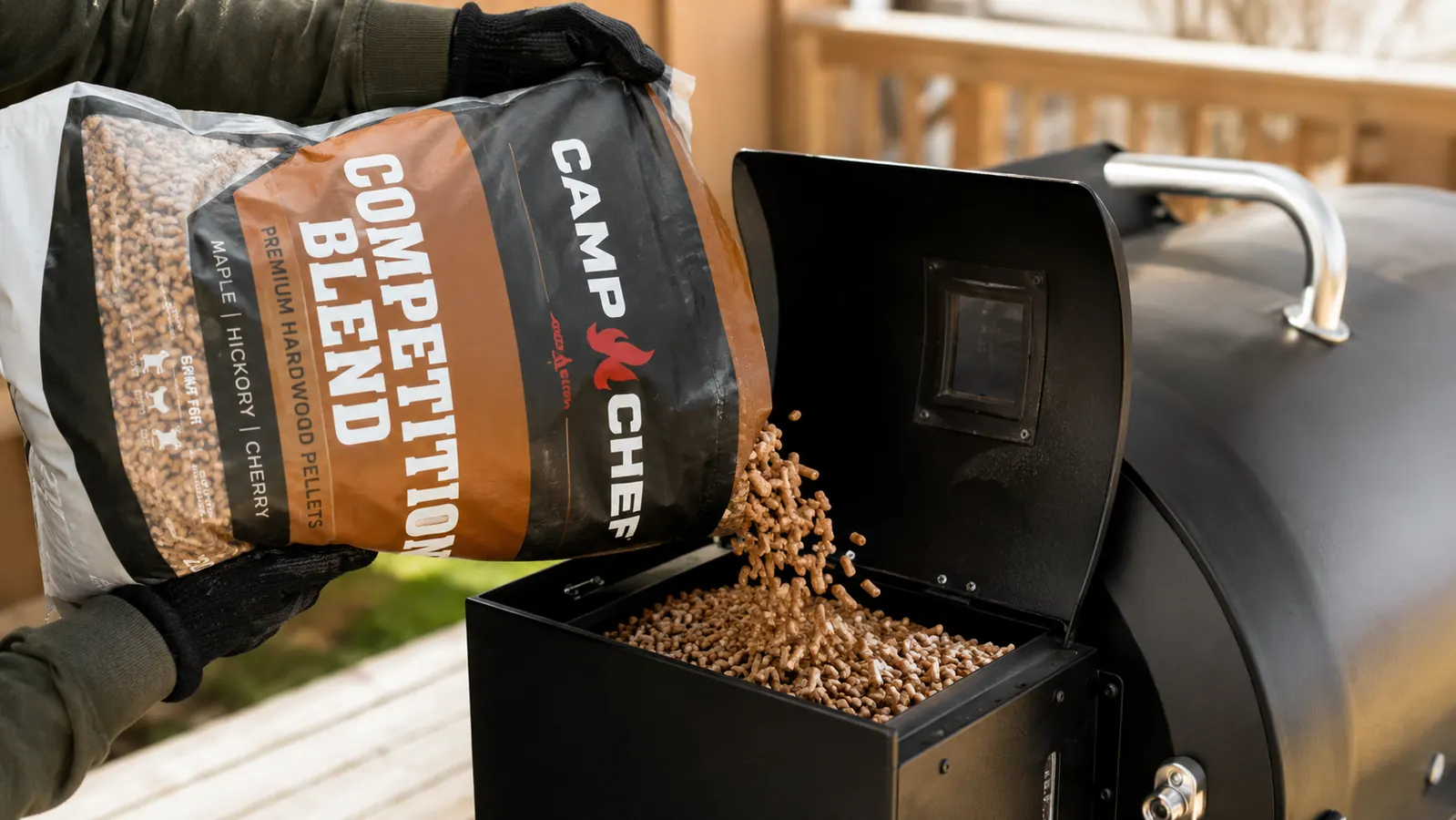 A hand pouring wood pellets from a bag into a smoker hopper, a few pellets scattering onto the top of the pile
