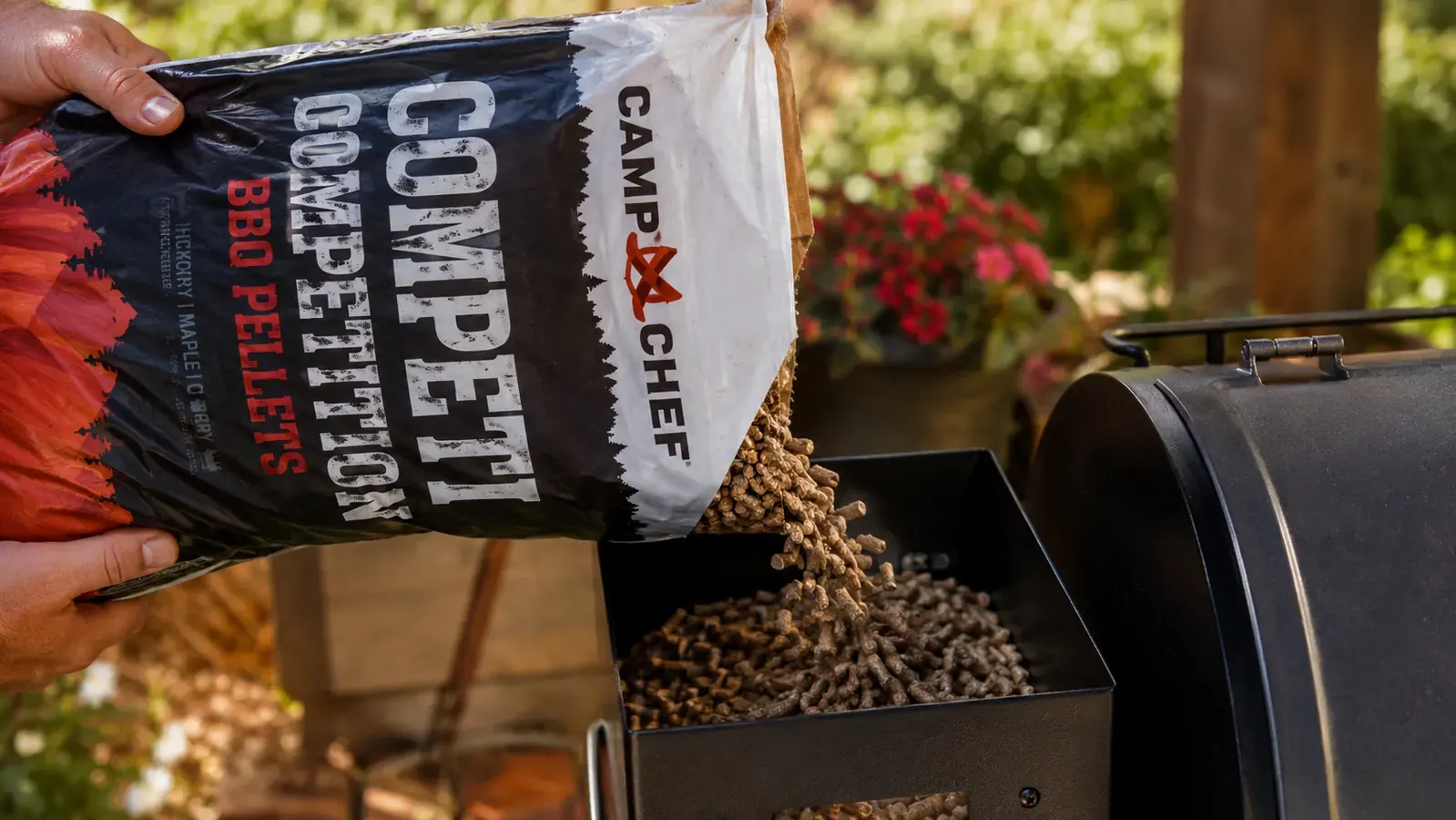 A hand pouring wood pellets from an open bag into the hopper of a smoker