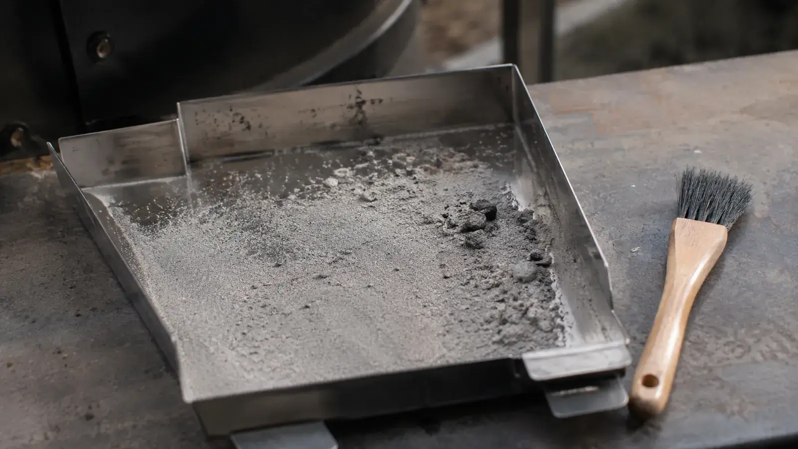 A stainless steel smoker ash tray dusted with a thin, light gray layer of burned pellet ash from a long cook