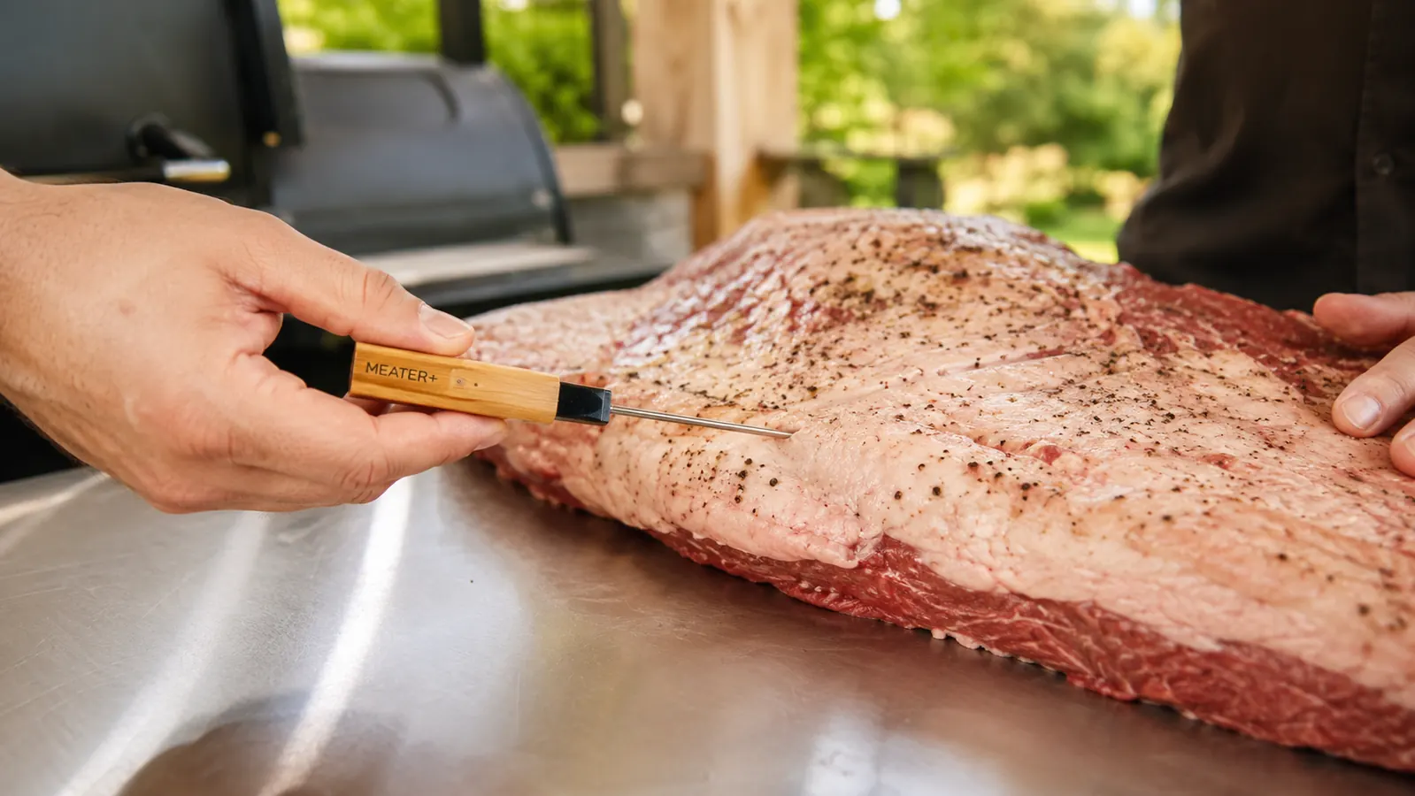 A hand lowering a thin meat probe into the flat of a brisket sitting on a stainless counter before it goes on the smoker