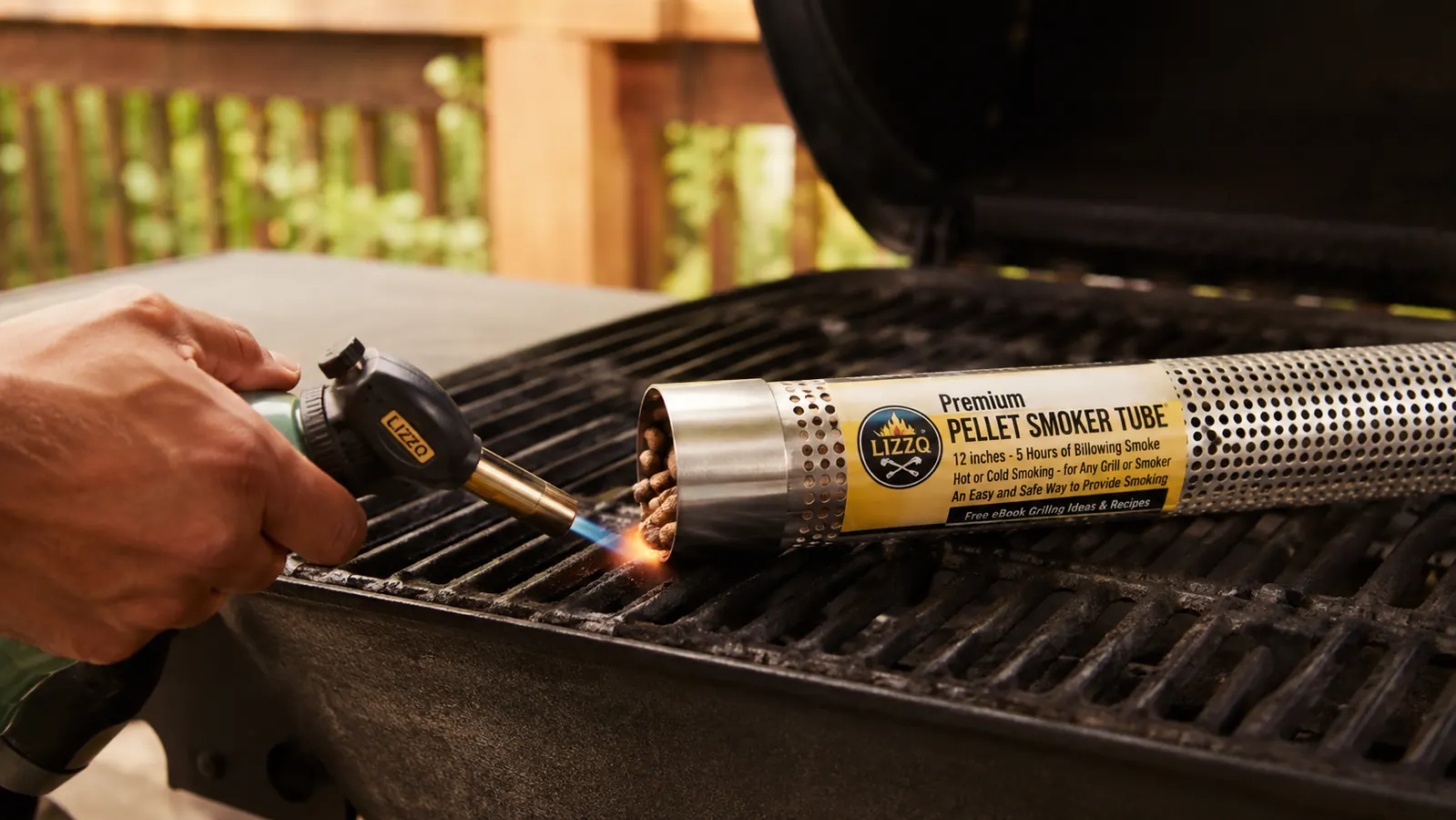 A hand holding a propane torch to the end of a metal smoke tube set on a grill grate, low flames just starting