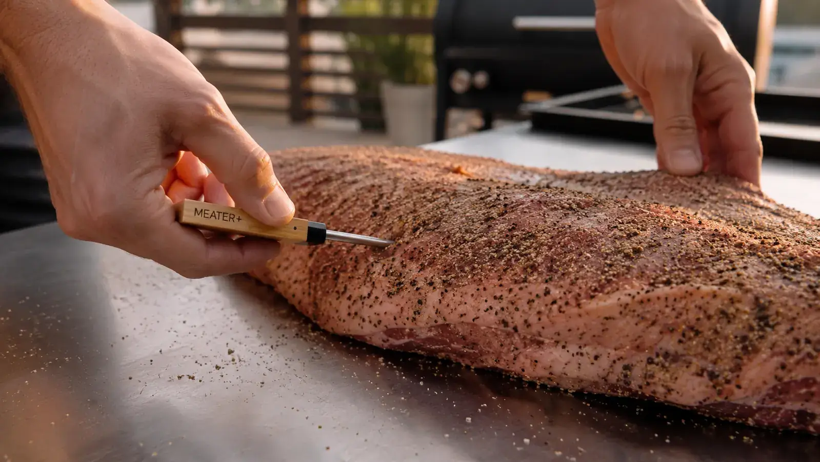 A hand sliding a long digital thermometer probe into a large trimmed brisket on a stainless work table