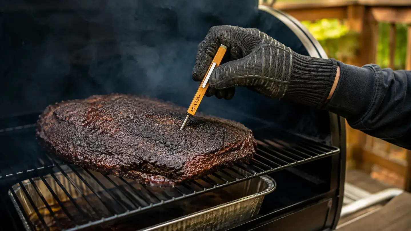 A hand with tongs checking a wire-free probe handle sticking out of a brisket on a smoker grate, stainless steel pan below
