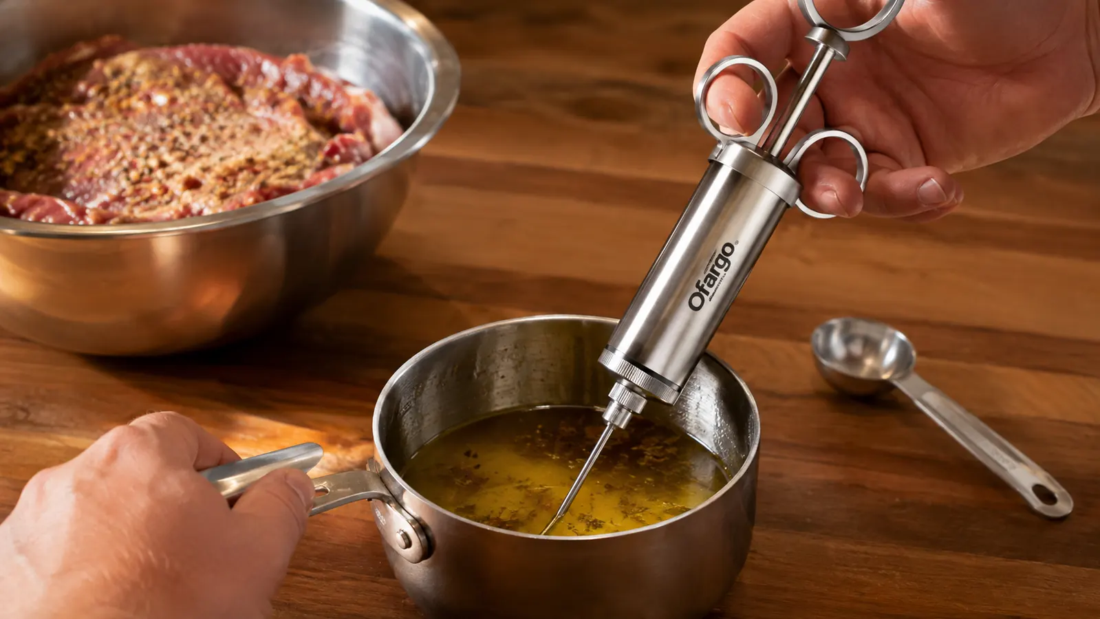 A hand filling a stainless steel meat injector from a small pot of warm marinade on a kitchen counter