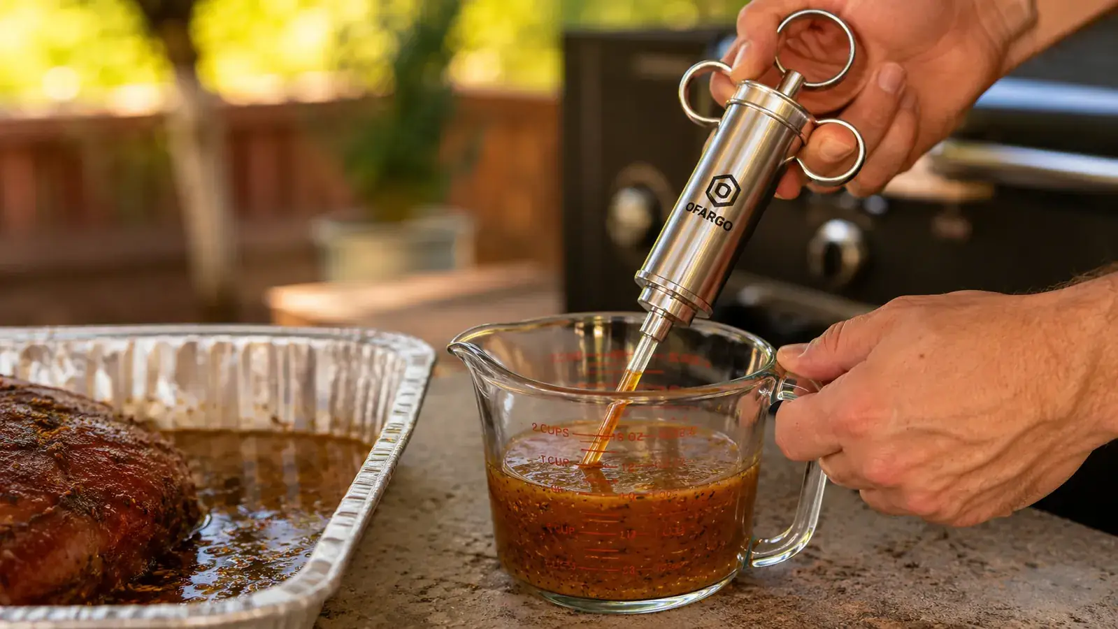 A man's hand filling a stainless injector barrel with warm marinade from a glass measuring cup beside a foil pan on a counter